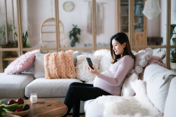 젊은 아시아 임산부는 아늑한 집에서 거실에서 소파에서 휴식을 취하고 스마트 폰을 사용하여 의사와 화상 통화 의료 약속을하고 있습니다. 기술, 원격 의학 및 임신 라이프 스타일 - smart phone china mobile phone asia 뉴스 사진 이미지