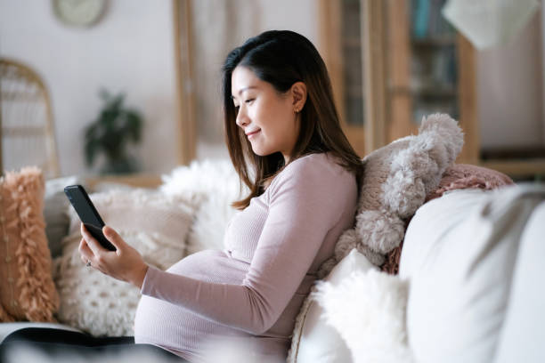 belle jeune femme enceinte asiatique utilisant un smartphone tout en se relaxant sur le canapé dans le salon, en profitant de la maison confortable. bien-être. mode de vie de grossesse - doctor asian ethnicity chinese ethnicity young adult photos et images de collection