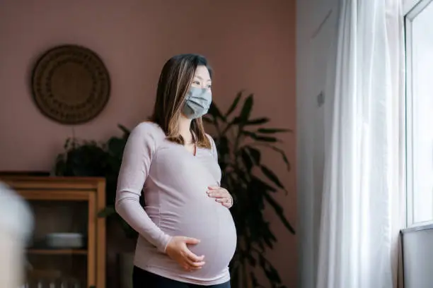 Portrait of young Asian pregnant woman with protective face mask to protect and prevent from the spread of virus. Standing by the window, cradling her baby bump, looking away thoughtfully in the living room at cozy home. Mother-to-be. Expecting a new life