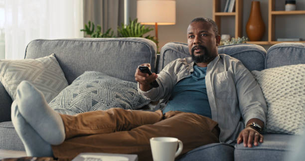 scatto di un uomo maturo che guarda la tv sul divano di casa - fare zapping foto e immagini stock