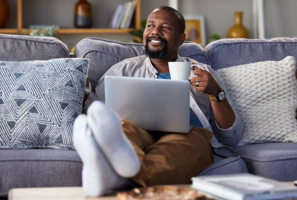 foto de un hombre maduro usando una computadora portátil y tomando café en el sofá de casa - banca electrónica fotografías e imágenes de stock