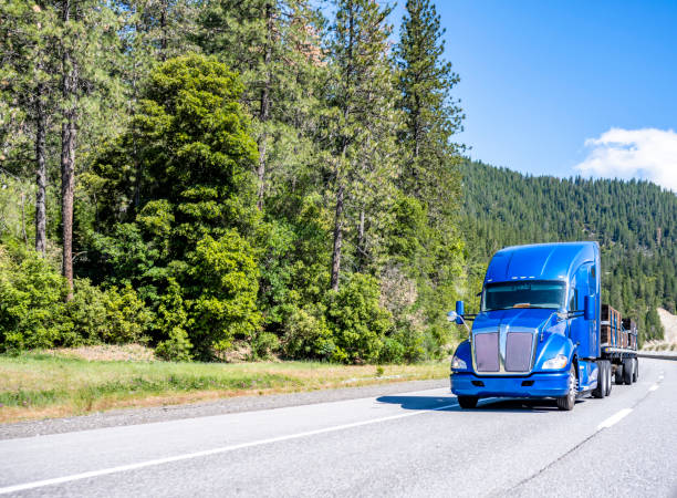 istock Bright shiny blue big rig semi truck tractor transporting heavy cargo on flat bed semi trailer climbing uphill on the road with forest on the side 1325693281