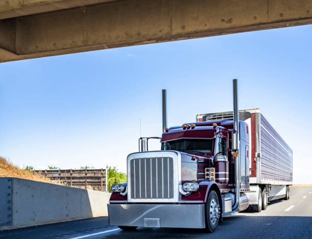 istock Classic big rig red semi truck tractor with vertical chrome pipes transporting cargo in refrigerated semi trailer running on the road under the bridge 1325695372