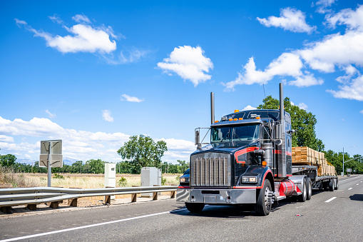 Classic black powerful big rig industrial semi truck tractor with red accents transporting lumber wood on step down semi trailer running on the divided highway road with meadow and trees on the side