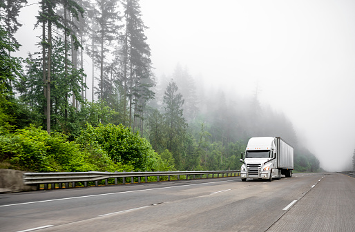 White big rig long haul semi truck transporting commercial cargo in dry van semi trailer running on turning one way highway road with forest on the hill at strong fog weather with poor visibility