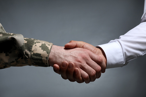 Soldier and businessman shaking hands against grey background, closeup