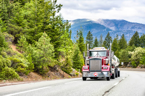 istock Powerful shiny red big rig classic bonnet semi truck with chrome accessories transporting fuel in stainless steel tank semi trailer running on the winding mountain road with green forest 1325699556