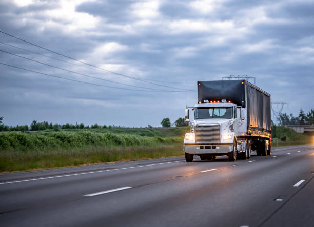 istock Old style classic white big rig day cab semi truck with turned on headlights transporting cargo in covered semi trailer running on the night highway road with light reflection 1325700631