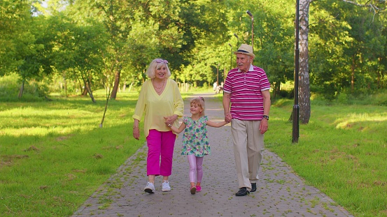Child girl kid walking with senior grandmother and grandfather. Cute granddaughter holding hands with her grandparents man and woman in park. Happy family relationship. Active life after retirement