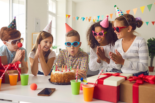 Birthday boy blows candles on a cake to the applause of his multiethnic friends or classmates at his party at home. Friends in colorful holiday accessories have fun in a festively decorated room.