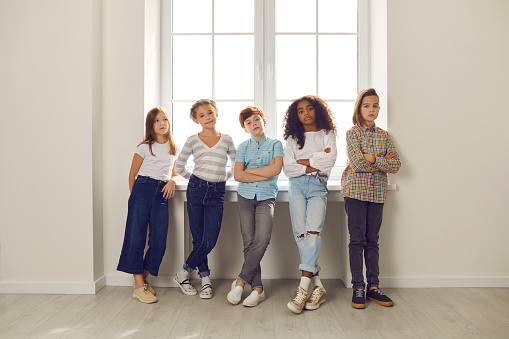 Group of serious diverse kids looking at camera standing by window with arms folded in hall at school or educational center. Success, confidence in future, conflict resolution in children's community