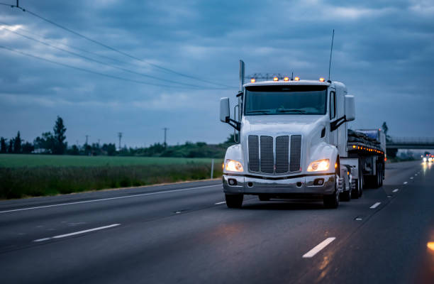 istock Low roof profile white big rig semi truck transporting fastened cargo on flat bed semi trailer with turned on side lights running on the night highway road 1325700961