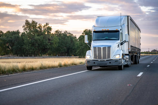 istock Powerful white big rig semi truck with turned on headlight transporting cargo in dry van semi trailer running on the evening highway road at twilight time 1325702016