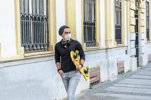 Young Student with Protective Face Mask is Carrying a Skateboard  During a Hot Summer Day in a Pandemic Time.