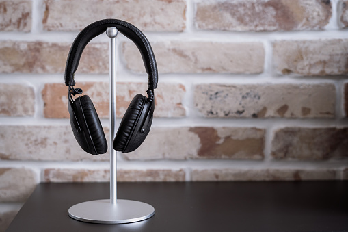 Close up of black wireless stereo headphones on white minimalistic stand against brick wall background on dark brown shelf surface. Modern audio equipment for listening music