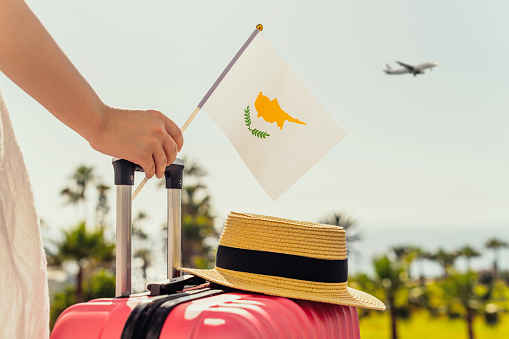 Woman with pink suitcase, hat and Cyprus flag standing on passengers ladder and getting out of airplane opposite sea coastline with palm trees