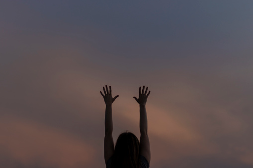 Woman holds hands raised up against the background of the evening sky.