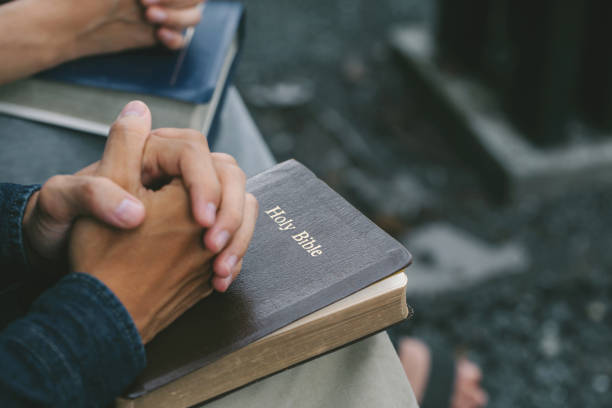 istock Hands folded praying over a Bible. Casual man praying with his hands together over a closed Bible, hands over soft-focus Bible. Holy Bible in church concept for faith, spirituality, worship, and religion. 1325724145