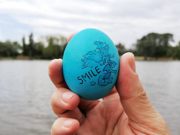 Decorated blue Easter egg with illustration and text smile in hand. The happiest Christian holiday. Smile, joy, happiness and love. The custom of tapping eggs. In the background river, forest and sky