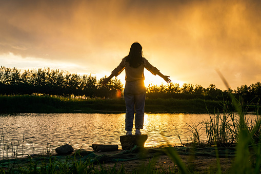 Young cheerful woman by the lake enjoying nature. Arms outstretched for positive emotion.