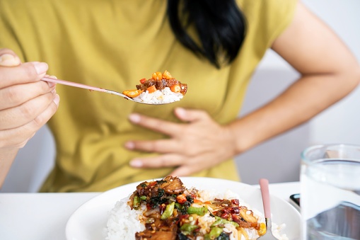 Mujer asiática comiendo comida picante y teniendo reflujo ácido o la mano de acidez estomacal sosteniendo una cuchara con chiles otra mano sosteniendo su estómago photo