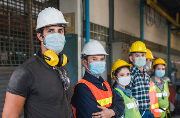 istock Group of diverse team of workers wearing face mask and protective helmets standing in front of the factory 1325751030