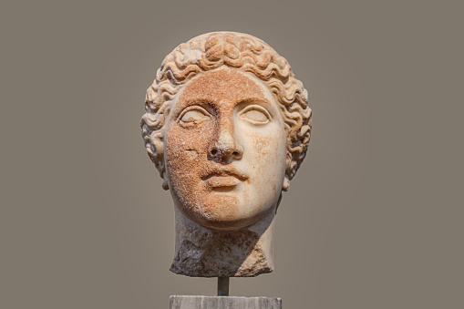 Damaged head of ancient Greek statue with part of face rough and orange from deterioration isolated against tan background.