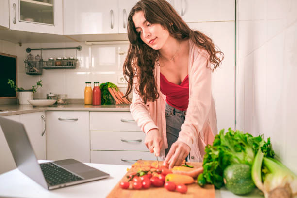 istock Young woman reading online recipes using laptop while cutting vegetables in the kitchen 1325764912