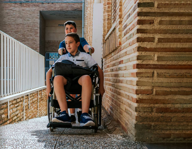 A boy pushing his friend's wheelchair up a ramp to enter school. Back to school and integration concept. stock photo