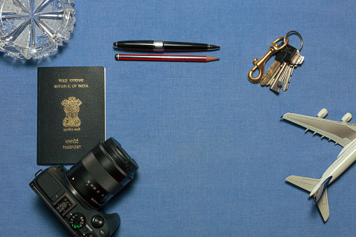 Ready to travel - A new India passport, a digital single lens reflex camera, a ballpoint pen, pencil and a bunch of keys indicating a desire to travel out of the country. The items are placed on a plain blue fabric. An airplane model has been placed at the bottom right. A crystal ashtray at the top left adds some pizzaz. The old Indian passport had the word, 