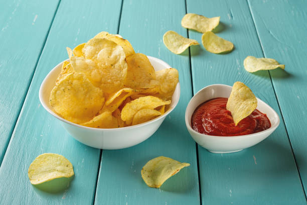 istock Delicious potato chips with dill and thick tomato ketchup in white ceramic bowls over blue wooden table. Popular crispy snack of potatoes deep-fried thin slices. Unhealthy eating and junk food. 1325798459