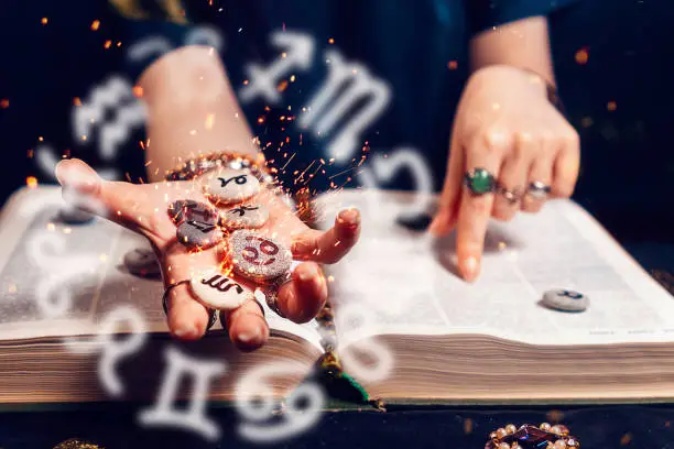 Astrology and horoscope. Fortune teller's hand holds the sparkling zodiac stones in the palm of her hand over the open book. The concept of divination and magic.