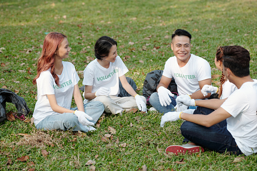 Cheerful young volunteers sitting on grass in circle, talking and laughing after collecting trash in city park or on campus