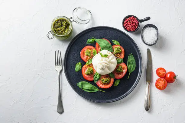 Buffalo burrata cheese served with fresh tomatoes and basil leaves pesto sauce on black plate white background space for text.