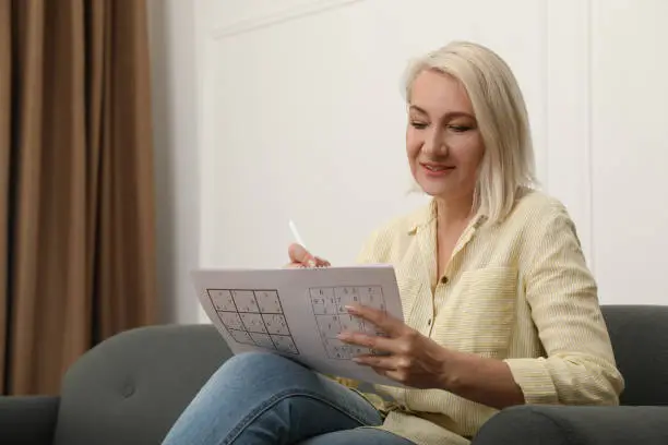 Middle aged woman solving sudoku puzzle on sofa at home