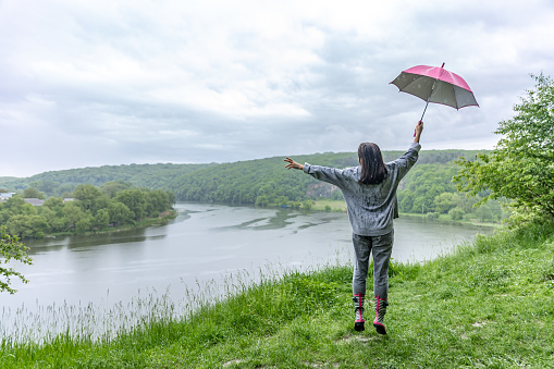 Back view of a girl under an umbrella jumping near a lake in a mountainous area in rainy weather.