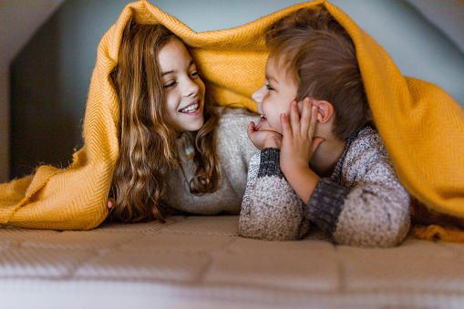 Happy little brother and sister talking while being covered with blanket on a bed. Copy space.