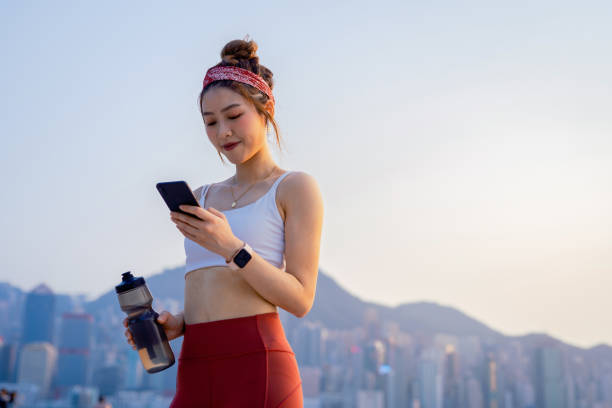 young asian sports woman using smartphone after exercising outdoors in city in the morning, against urban city skyline. relaxing after running. youth culture. habits and hobbies. active lifestyle. health and fitness with technology concept - self improvement audio imagens e fotografias de stock