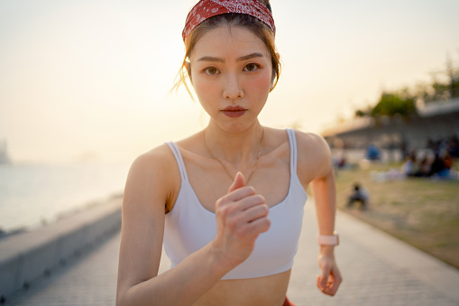 Portrait of determined young Asian sports woman exercising outdoors, running in urban park by the promenade in city at sunset. Active lifestyle. Health and fitness concept