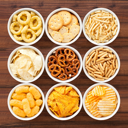 Top view of nine bowls containing varieties of salty snacks