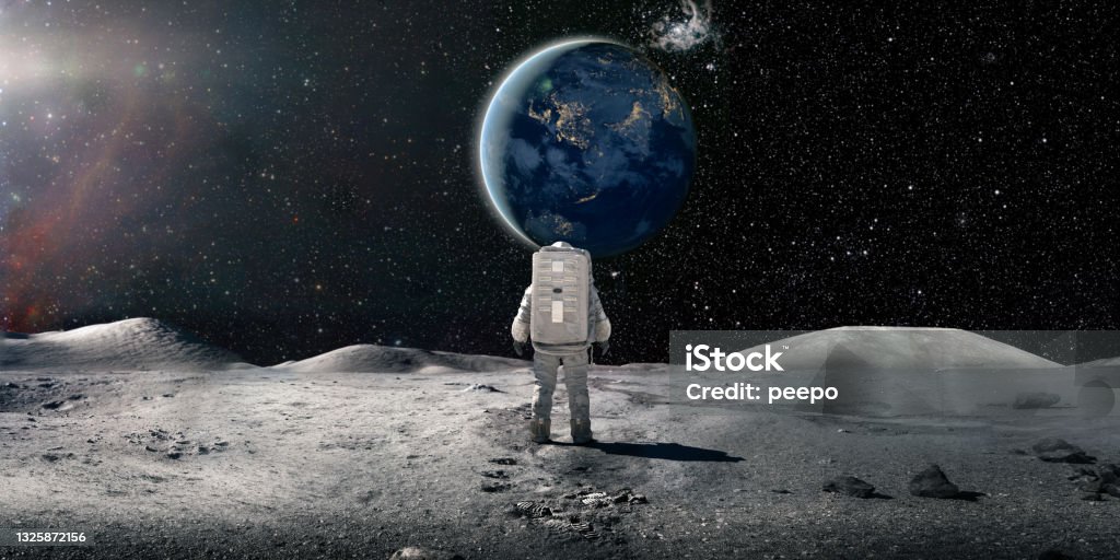 Lone Astronaut In Spacesuit Standing On The Moon Looking At The Distant Earth A single astronaut viewed from behind, standing and looking at the sun rising over a distant plant earth surrounded by distant stars and galaxies. The astronaut is wearing a conventional white spacesuit with backpack. Earth image from NASA: https://eoimages.gsfc.nasa.gov/images/imagerecords/79000/79790/city_lights_asia_night_8k.tif Moon Surface Stock Photo