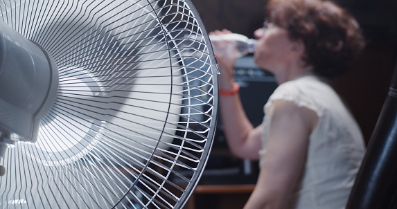 Woman is trying to work at the desktop computer at home, suffering from hot weather and drinking water while using electric fan