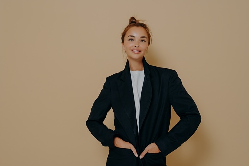 Stylish caucasian business woman in black suit with hair tied up in high bun and with light makeup posing on beige background in studio. Fashion style body length studio portrait. Career concept