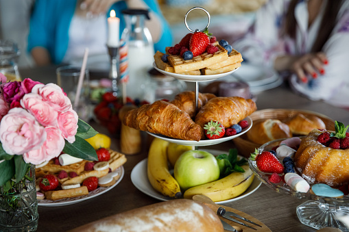 Arrangement of delicious looking sweet food such as waffles, croissants, a cake and bunch of fresh fruit as decoration, served on cake stands on a dining table to be enjoyed during afternoon tea.