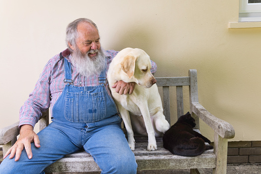 Portrait of a smiling old man sitting in his garden with his dog. Old man relaxing outside with his dog and both looking at the camera. Photo of a happy old man having a good time with his dog in his garden.