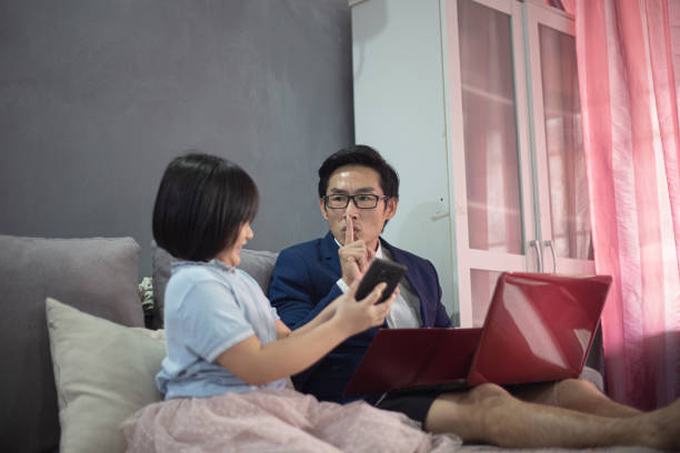 アジアの中国人男性は、オンライン会議中にソファに座っているラップトップを使用して、短いパンツと適切なスーツで自宅で働いています。彼が会っている間、彼の子供たちは不安を感じ� - inconvenience meeting business distracted ストックフォトと画像