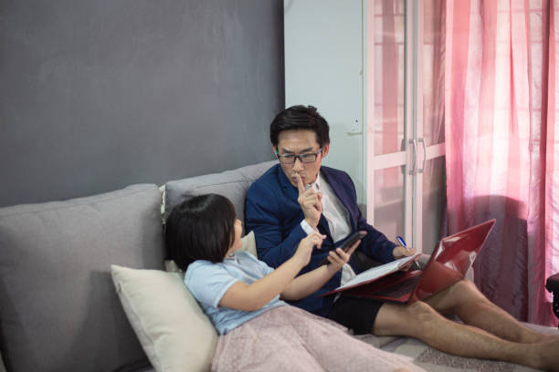アジアの中国人男性は、オンライン会議中にソファに座っているラップトップを使用して、短いパンツと適切なスーツで自宅で働いています。彼が会っている間、彼の子供たちは不安を感じ� - inconvenience meeting business distracted ストックフォトと画像