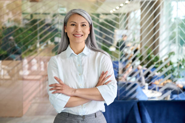 happy smiling confident middle aged asian older senior female leader businesswoman standing in modern office workplace looking at camera arms crossed. business successful executive concept. portrait. - mid aged woman imagens e fotografias de stock