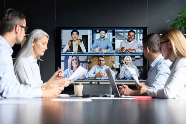 Photo of Global corporation online videoconference in meeting room with diverse people sitting in modern office and multicultural multiethnic colleagues on big screen monitor. Business technologies concept.