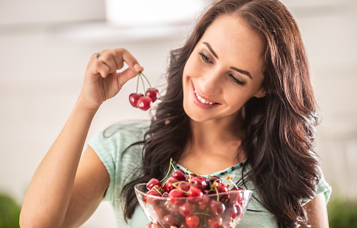 Good-looking girls looks at several cherries in her hand while holding bowl of them in the other one.